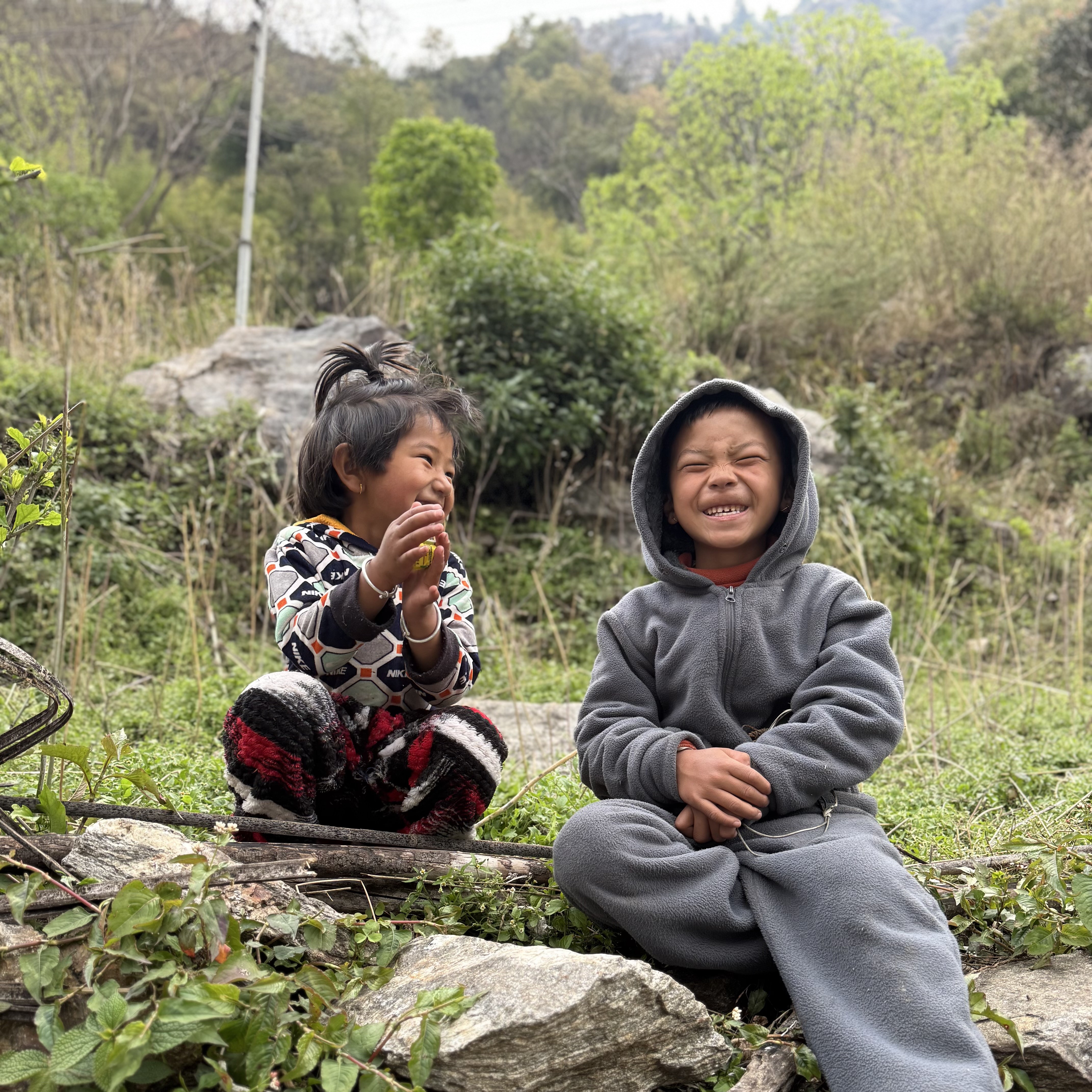 Two children sitting on a fallen log beside the trail, both laughing, the younger one holding a sweet