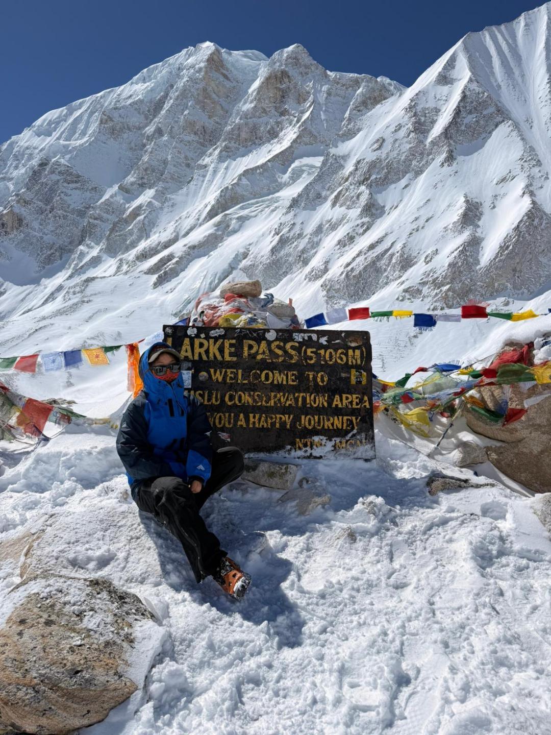 Dolkar beside the Larke Pass signboard at 5,106 m, prayer flags and deep snow, the mountain face above