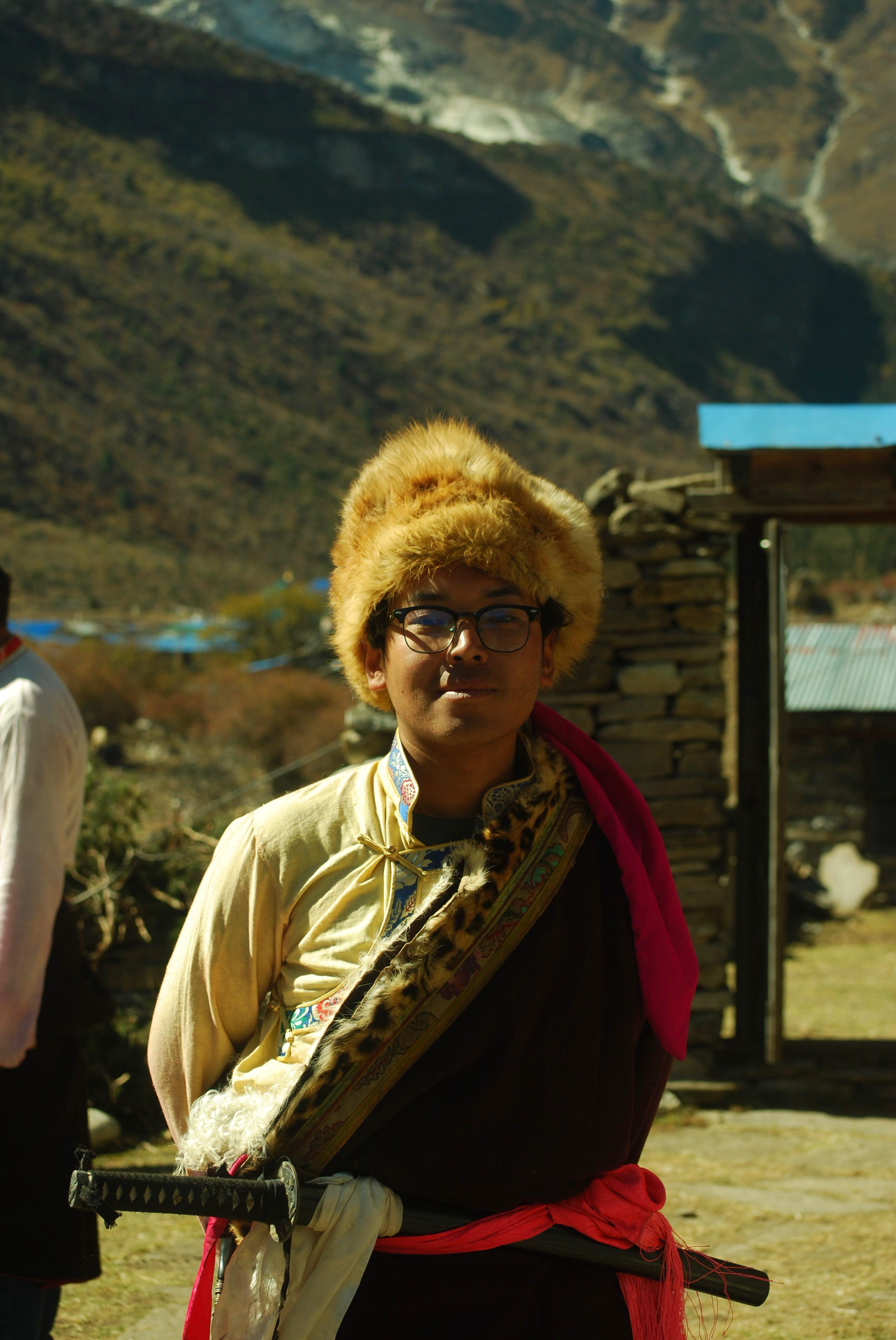 Jigme Dorje in traditional Nubri dress, a sword at his red sash, standing by a stone wall below the valley slopes
