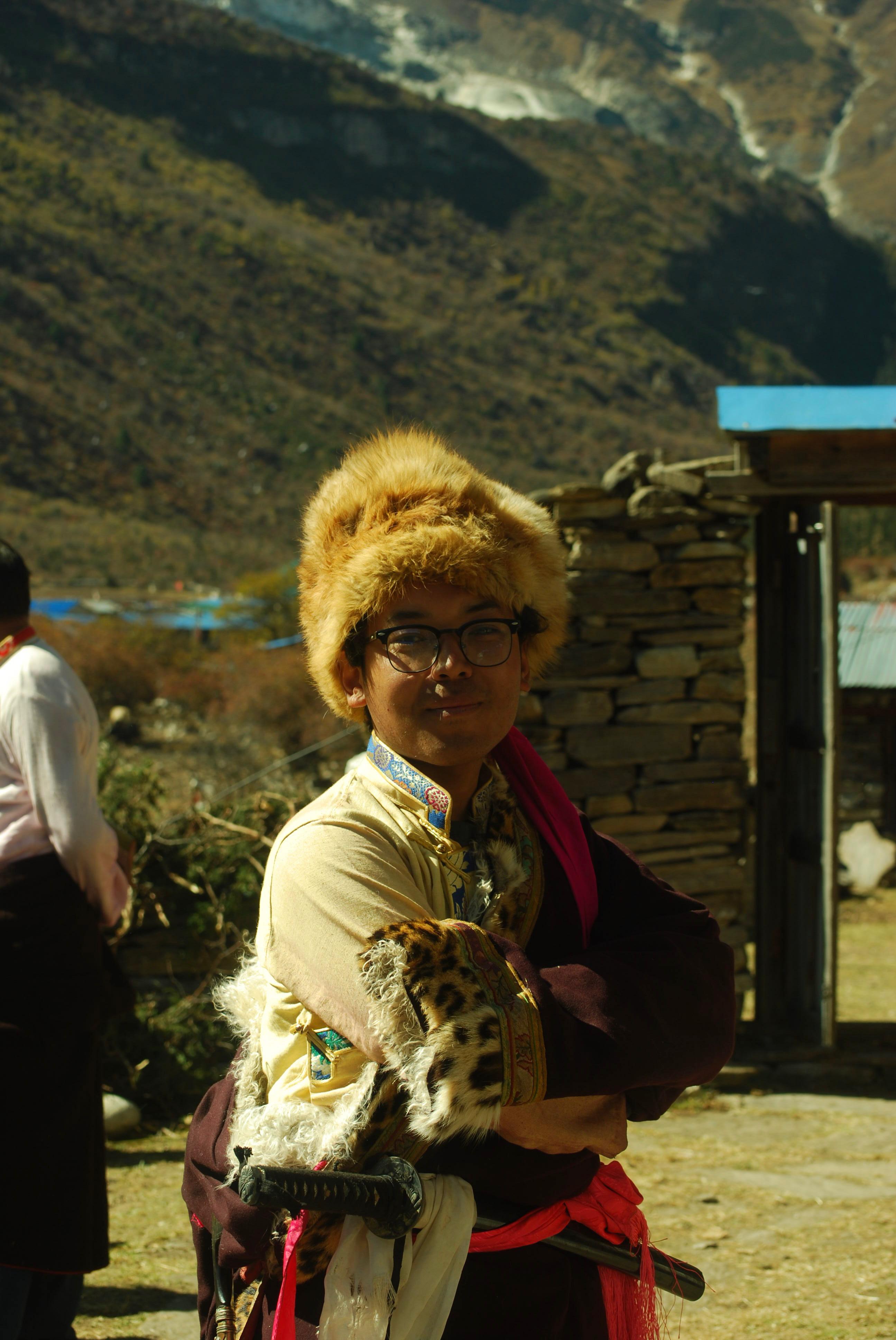 Jigme Dorje, founder of Drokpo, in traditional Nubri dress — golden fur hat, brocade collar and leopard-trimmed chuba — arms folded in his village, the valley wall behind him